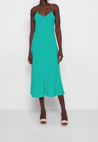 Femme portant une robe midi turquoise sans manches avec des bretelles spaghetti et des talons ouverts beiges, se tenant devant un fond uni.