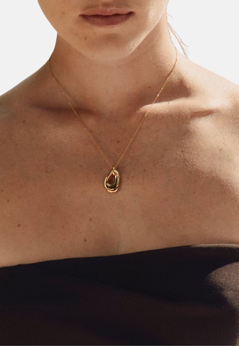 Primer plano de una persona que lleva un collar de cadena dorada con un colgante abstracto entrelazado sobre hombros descubiertos y un top negro sin tirantes.