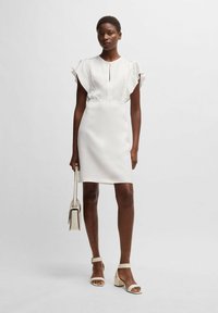 Biała sukienka z krótkimi falbaniastymi rękawami, dekoltem w kształcie łezki, dopasowaną talią i prostą spódnicą. W połączeniu z beżowymi sandałami na słupku i mini torebką.