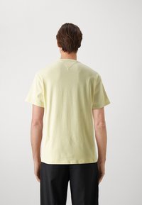 Yellow cotton t-shirt with short sleeves, a rounded neckline, and a small V-shaped detail at the back, paired with dark pants.