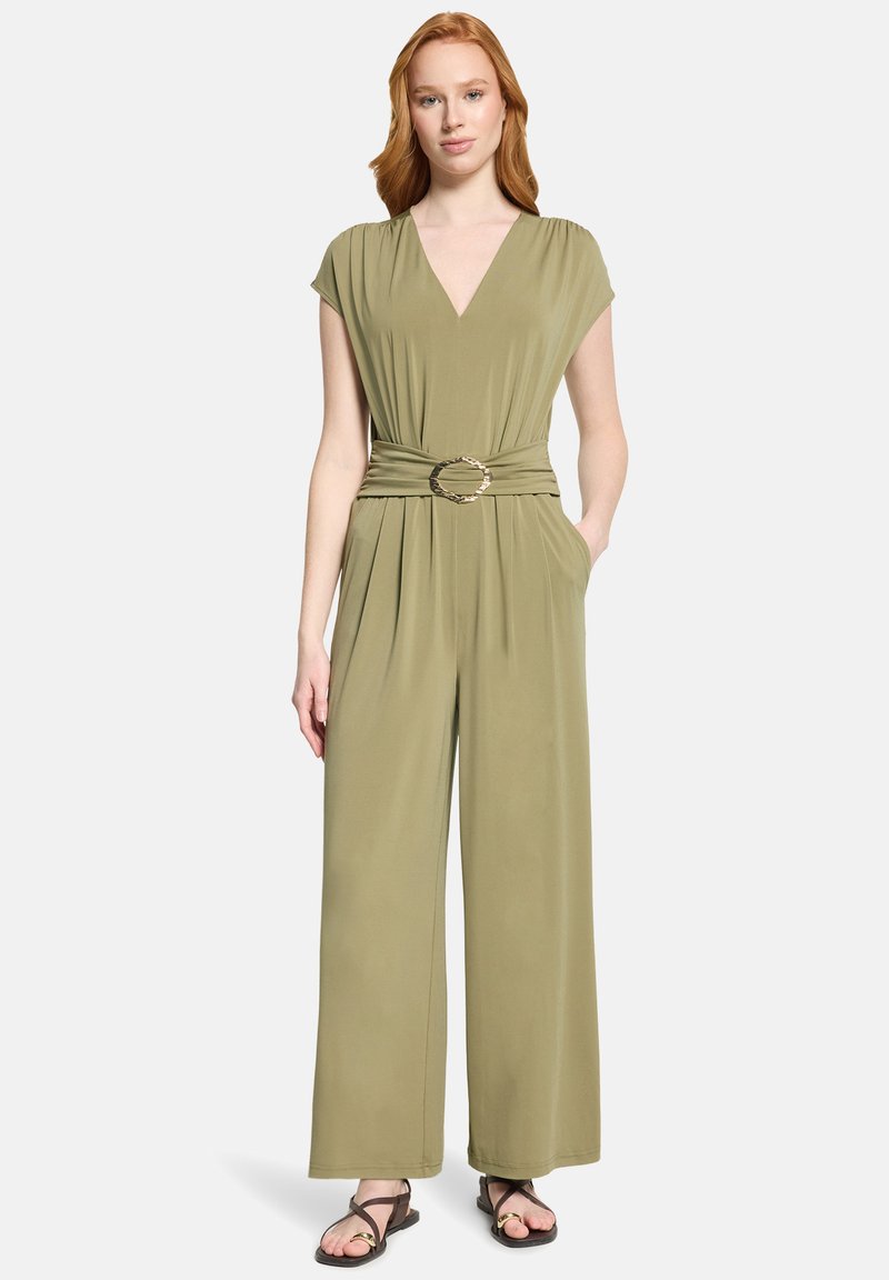Femme portant une combinaison pantalon kaki à jambes larges avec un décolleté en V, des manches courtes, une taille cintrée par une ceinture à boucle circulaire et des sandales noires.