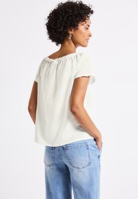 Top beige chiaro a maniche corte con scollo elasticizzato, caratterizzato da una vestibilità comoda e un tessuto morbido e texturizzato, abbinato a jeans blu.