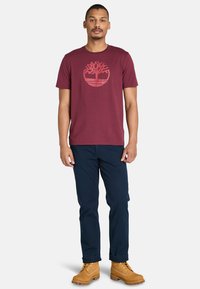 Burgundy bomullströja med en röd träddesign, kombinerad med marinblå byxor och bruna läderstövlar med snörning och en robust sula.