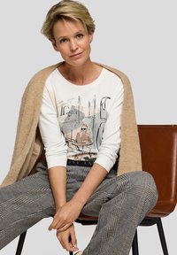Frau mit kurzen blonden Haaren, die einen beigen Mantel über einem weißen Langarmshirt mit Grafik und Karohose trägt, sitzt auf einem braunen Stuhl vor einem grauen Hintergrund.