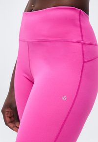 Κοντινή λήψη της μέσης και του μηρού ενός ατόμου που φορά φωτεινό ροζ αδιάκοπο κολάν με ορατή ραφή και μικρό αντανακλαστικό λογότυπο της μάρκας.