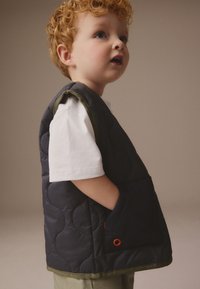 Vestă matlasată bleumarin cu margini verzi și un buzunar în față, purtată peste o cămașă albă. Prezintă un accent rotund portocaliu pe buzunar.