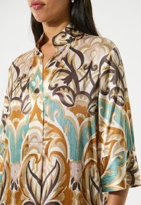 Blouse en soie avec un col haut et des manches évasées. Présente des motifs floraux complexes dans des teintes de marron, bleu-vert et crème. Texture légèrement brillante.