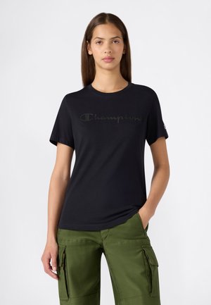 Junge Frau mit langen Haaren, die ein schwarzes Champion-T-Shirt und grüne Cargohosen trägt, steht mit einer Hand in der Tasche vor weißem Hintergrund.