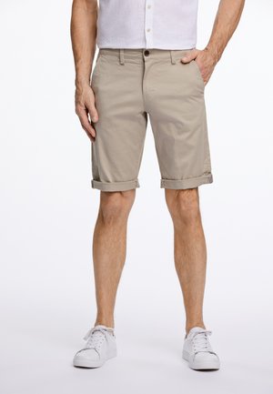 Beige bomuldsshorts med oprullede manchetter, forlommer og knaplukning. Modellens iført hvide sneakers og en lysfarvet skjorte.