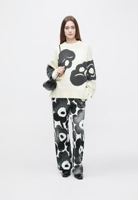 Per didelį kūno dydžio kreminis megztinis su juodu gėlių raštu, derinamas su plačiais juodai baltais gėlių raštu kelnėmis. Modelis laiko mažą juodą rankinę.