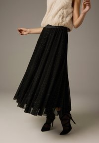 Zwarte geplooide maxi-rok van doorschijnende stof, versierd met zilveren spikkels. Model draagt een beige top met structuur en zwarte schoenen met hak.