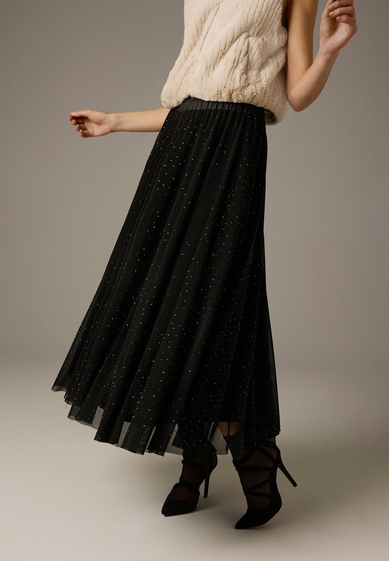 Zwarte geplooide maxi-rok van doorschijnende stof, versierd met zilveren spikkels. Model draagt een beige top met structuur en zwarte schoenen met hak.