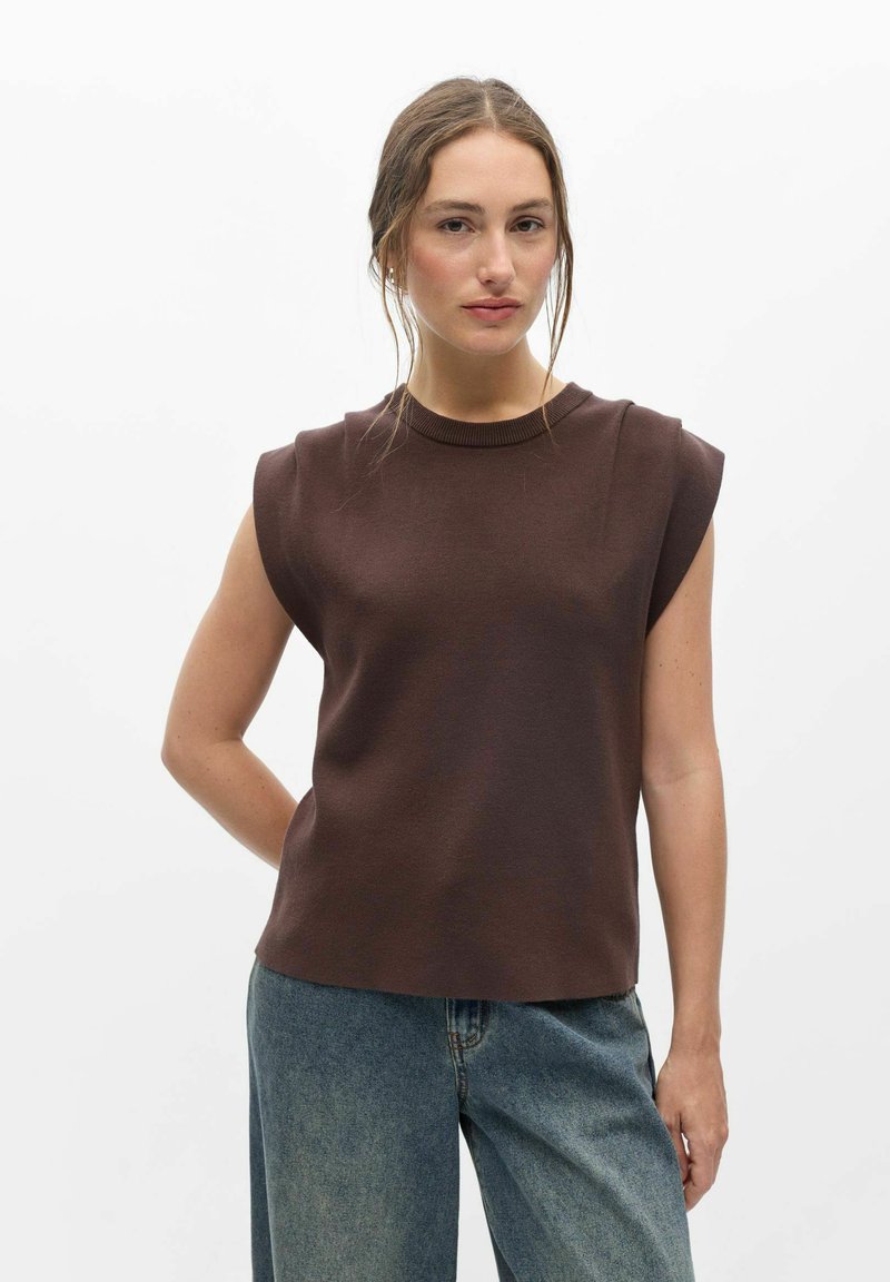 Jeune femme portant un haut marron sans manches et un jean bleu, debout sur un fond blanc uni, regardant la caméra avec une expression neutre.