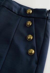 Laivastonsiniset housut, joissa on sileä kangas, neljä koristeellista kultanappia ja räätälöity malli sivussa olevalla vetoketjulla.