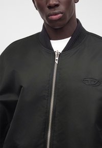 Gros plan d'une personne portant une veste noire zippée avec un col côtelé et un logo brodé sur la poitrine gauche.