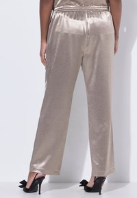 Donna che indossa pantaloni larghi beige lucidi abbinati a tacchi alti neri con grandi fiocchi, in piedi contro un muro bianco.