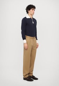 Tummansininen pitkähihaisempi polo valkoisella kauluksella, vaaleat laskostetut housut, mustat loafers-kengät kultaisin yksityiskohdin. Malli seisoo yksinkertaista taustaa vasten.