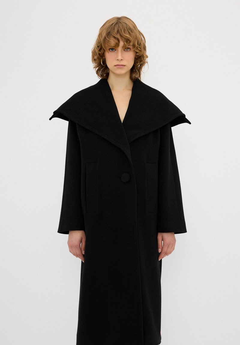 Cappotto di lana nero con un drammatico colletto oversize, due tasche laterali e chiusura a un bottone. Tessuto morbido, design fino al ginocchio.