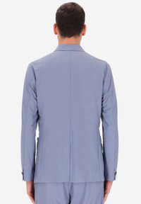 Uomo con un giacca di suit blu chiaro e pantaloni coordinati, mostrato di spalle su uno sfondo bianco semplice.