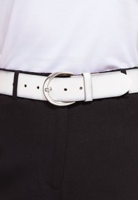 Cintura bianca con fibbia argentata fissata attorno a pantaloni neri e camicia bianca infilata.