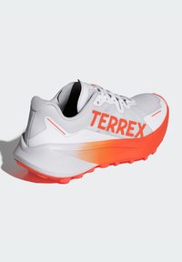 Scarpa da ginnastica bianca con una suola a gradiente arancione e il testo "TERREX". Presenta una tomaia in mesh traspirante e dettagli testurizzati.
