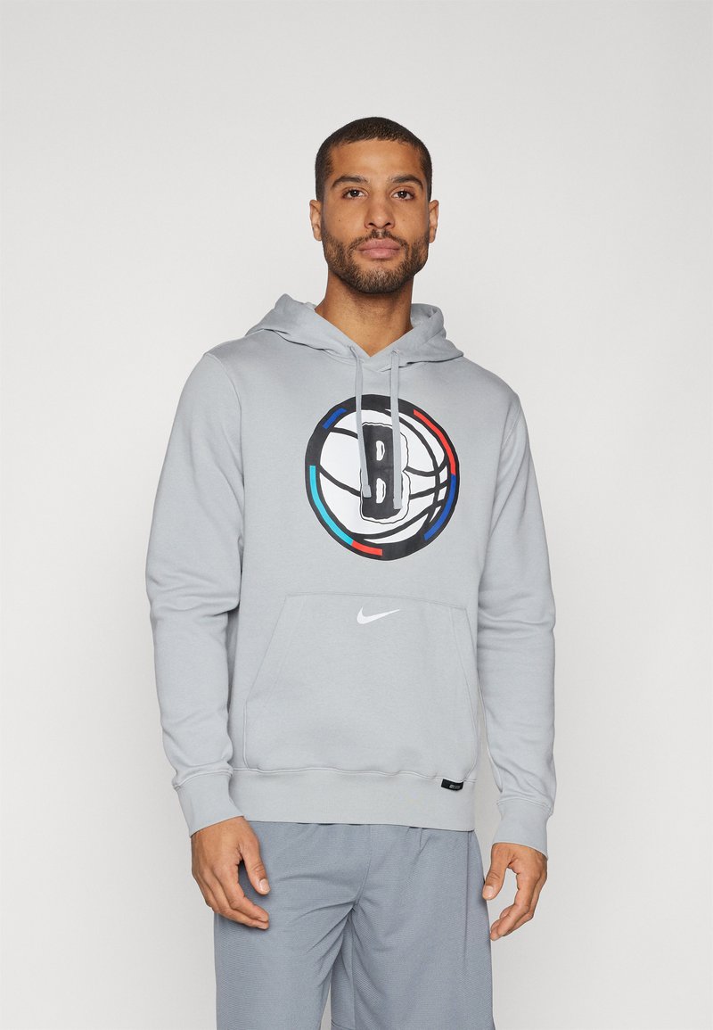 Pilkas džemperis su gobtuvu, su centriniu krepšinio piešiniu, kuriame yra ryškus „B“ logotipas, kengūros tipo kišenė, su raišteliais gobtuvas ir mažas „Nike“ logotipas juosmens srityje.