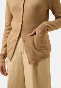Lichtbruin wollen cardigan met een knoopsluiting aan de voorkant, voorzien van twee voorzakken en geribbelde manchetten, gecombineerd met beige ruime broek.