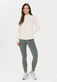 Hellbeige Zip-Jacke mit hohem Kragen, kombiniert mit engen dunkelgrünen Leggings und grauen Sportschuhen. Glattes Material, minimalistisches Design.