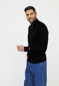 Pull en tricot noir avec un col haut et une fermeture éclair quart, présentant des poignets et un ourlet côtelés. Inclut un accent marron sur le poignet. Porté avec un jean bleu.