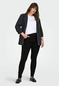 Fekete szabott blézer, fehér mintás rövid ujjú ing, fekete slim fit farmersz nadrág és fekete lapos cipők. A modern, laza dizájnra helyezve a hangsúlyt.