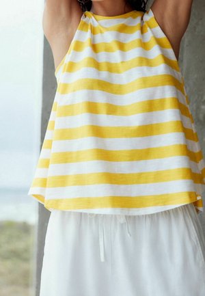 Femme portant un haut sans manches à rayures jaunes et blanches avec un pantalon blanc ample, levant les bras au-dessus de sa tête à l'intérieur près d'une fenêtre.