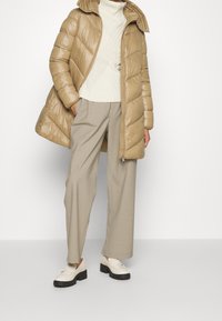 Giacca puffer beige con motivo trapuntato, abbinata a un maglione in knit crema, pantaloni grigio chiaro e mocassini bianchi con suole nere.