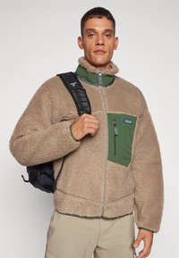 Brown fleece jacket with a high collar, green zippered pocket, and gray accents, worn with a black backpack over a light-colored outfit.