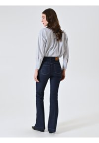 Donna con lunghi capelli castani che indossa una camicia a righe con bottoni e jeans bootcut blu scuro, in piedi e che guarda di lato.