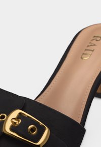Mule noir avec un détail de boucle dorée et une semelle intérieure beige estampillée "RAID" en lettres dorées.