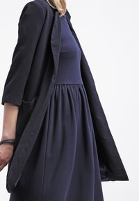 Robe bleu marine à jupe évasée et taille froncée, superposée sous une veste assortie à devant ouvert. Le tissu semble doux avec une texture subtile.