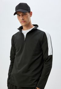 Sudadera negra de media cremallera con acentos blancos en los hombros, fabricada en un tejido suave; combinada con una gorra negra, diseño ajustado y mangas largas.