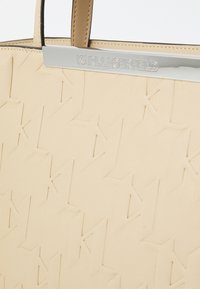 Beige taška s embosovaným logom a striebornými doplnkami. Hladký povrch a štruktúrovaný tvar s dvojitými hornými uchami.