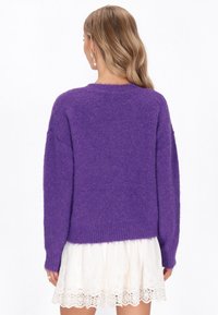 Fioletowy, puszysty sweter w oversize'owym stylu z długimi rękawami i ściągaczem, w zestawie z białą spódnicą wykończoną koronką. Brak widocznych ozdób lub akcentów.