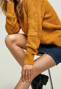 Senfgelber gestrickter Pullover mit Diamant- und Wellenmustern, kombiniert mit Jeansshorts und schwarzen Knöchelstiefeln auf einem silbernen Hocker.