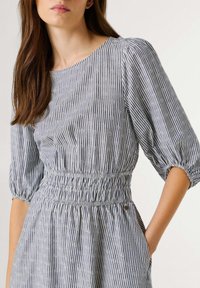 Žena u plavoj i bijeloj haljini s okomitim prugama i sitnim cvjetnim uzorkom, elastičnim strukom i napuhanim rukavima do tri četvrtine.