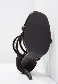 Chaussure à talon haut en daim noir avec fines brides et bout pointu. Elle présente une semelle noire lisse avec une texture antidérapante et un accent logo doré.