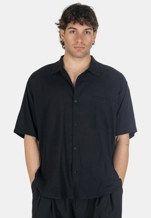 Giovane uomo con capelli ricci castani indossa una camicia nera a maniche corte, larga, con taschino sul petto, mani nelle tasche, espressione neutra, sfondo bianco.