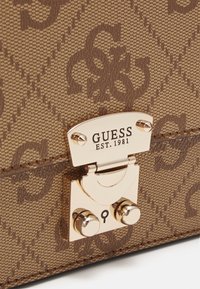 Sac en tissu marron à motifs avec un fermoir en métal doré gravé "GUESS EST. 1981" et un symbole de serrure sur la plaque de verrouillage.