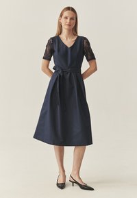 Tumesinine kleit on lühikeste pitsvarrukatega, V-kaelusega ja vööga. Seelik on lehvikukujuline ja lõpeb põlve all. Sobitatud mustade slingback kontsadega.