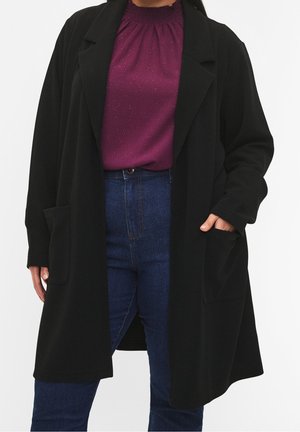Femme portant un long manteau noir sur un chemisier violet scintillant et un jean bleu taille haute, avec une main dans la poche du manteau.