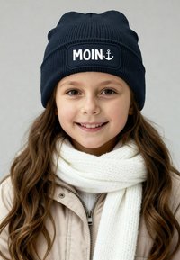 Navy knitted beanie with a white "MOIN" text and anchor design. Paired with a white textured scarf and a light beige jacket.