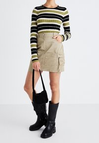 Gestreepte ribbel top in zwart, groen en wit; beige cargorok met zijzakken; zwarte kniehoge laarzen; zwarte franjehandtas.