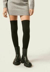Czarne prążkowane skarpetki do kolan zestawione z masywnymi czarnymi skórzanymi botkami za kostkę, charakteryzującymi się eleganckim designem i solidnymi teksturowanymi podeszwami.