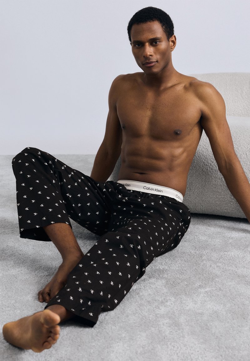 Μαύρο παντελόνι πιτζάμας με λευκό σχέδιο και ελαστική μέση με το λογότυπο "Calvin Klein", φοράει ένας καθιστός ανδρικός μοντέλος γυμνού κορμού.
